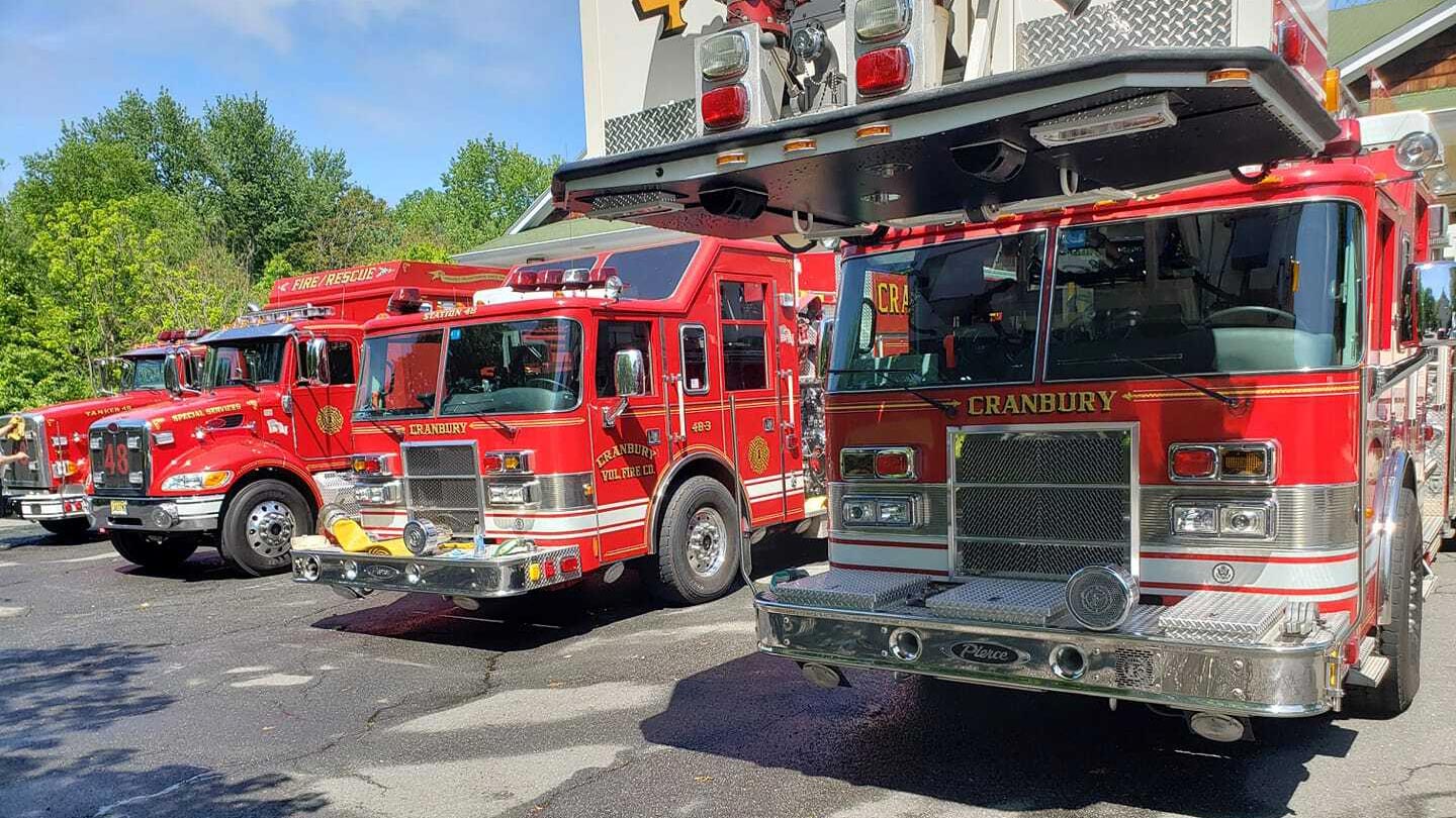 Three bright red fire trucks from Cranbury Fire Department are parked side by side outdoors on a sunny day, with trees and a building visible in the background.