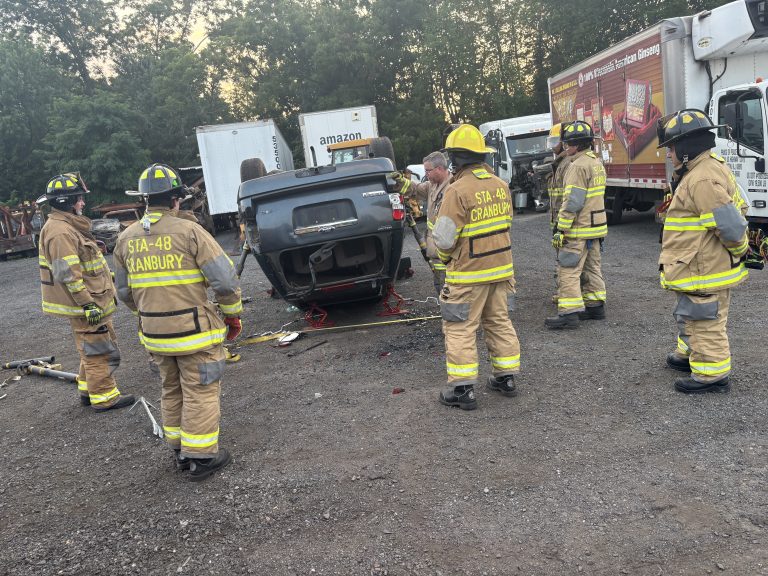 Six firefighters in yellow gear stand around an overturned vehicle in a gravel lot, with tools on the ground and trucks, including an Amazon truck, in the background. Trees are visible behind the vehicles.