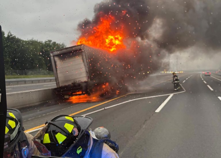 A large truck on a freeway is engulfed in flames and thick black smoke, while firefighters in helmets approach the scene. Rain droplets are visible on the camera lens, and traffic is light on the wet highway.