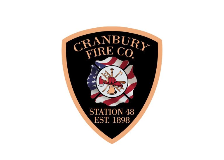 A black and gold shield-shaped emblem reads "Cranbury Fire Co. Station 48 Est. 1898" with a firefighter helmet, ladder, axe, and hose over a waving American flag background in the center.