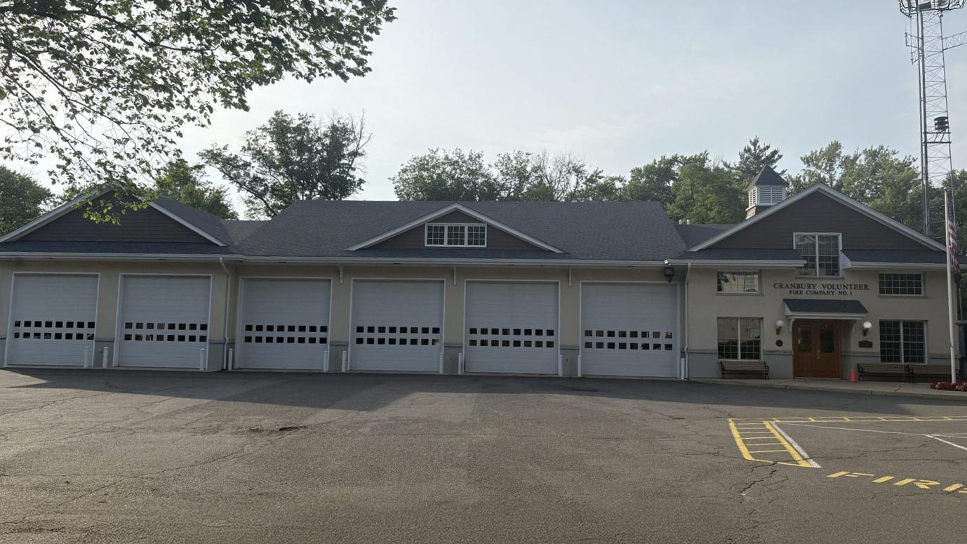 A large fire station building with six closed white garage doors, a gray roof, and a sign reading "CRANBERRY VOLUNTEER FIRE DEPARTMENT" above the entrance on the right side. Trees are visible in the background.