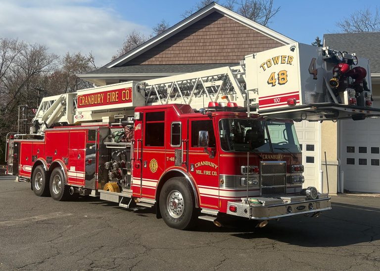 A large red fire truck with an extended ladder, labeled "CRANBURY FIRE CO" and "TOWER 48," is parked outside a building with white garage doors on a sunny day.