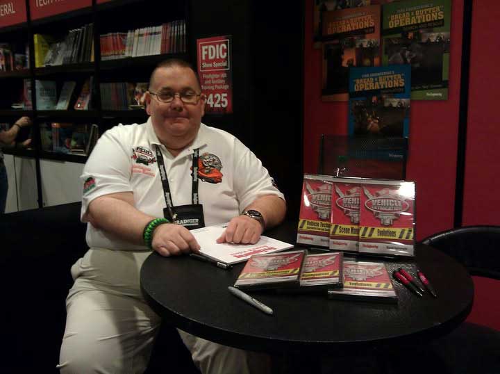 A man in a white shirt sits at a table with DVDs, pens, and brochures at an event booth. Shelves with books and posters are visible in the background. He is smiling and looking at the camera.