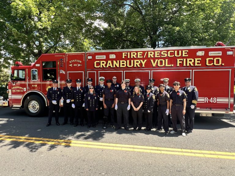 A group of firefighters in formal uniforms stands in front of a red Cranbury Volunteer Fire Company fire/rescue truck, parked on a sunny street with green trees in the background.