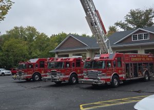 Four red fire trucks are parked outside a building, with one truck's tall ladder extended upward. The ground has yellow "FIRE" and "FIGHT" markings. Trees and cloudy sky are visible in the background.