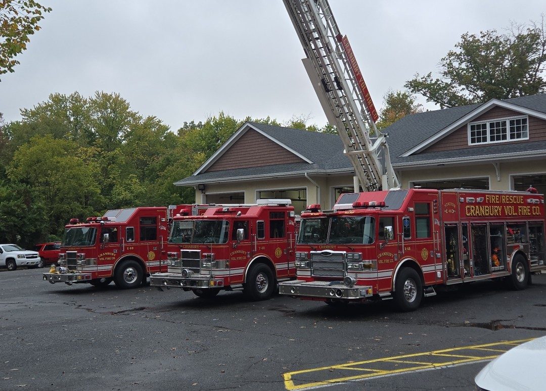 Four red fire trucks are parked outside a building, with one truck's tall ladder extended upward. The ground has yellow "FIRE" and "FIGHT" markings. Trees and cloudy sky are visible in the background.