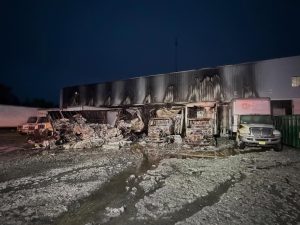 A warehouse at night with severe fire damage; the building’s exterior and trucks parked outside are charred, debris is scattered, and two trucks are visibly burned while others remain mostly intact.