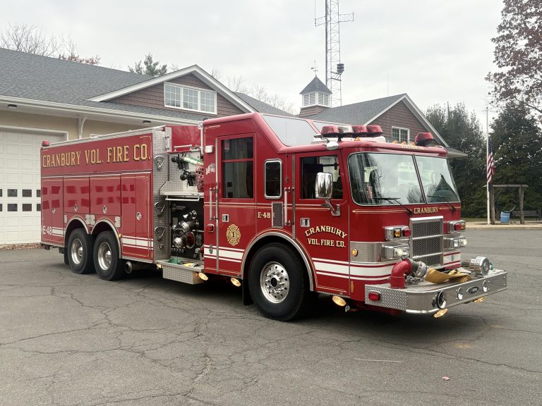 A red Granbury Volunteer Fire Company fire truck is parked outside a fire station on a paved lot, with buildings, trees, and an American flag in the background.